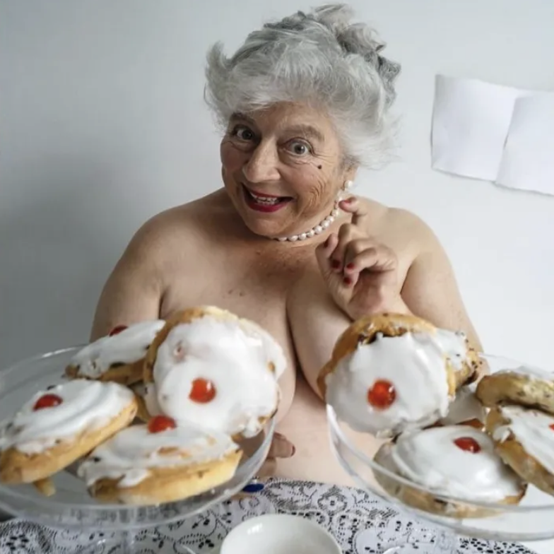 An elderly woman with gray hair smiles and poses playfully while holding two glass plates of homemade cookies topped with icing and cherries.