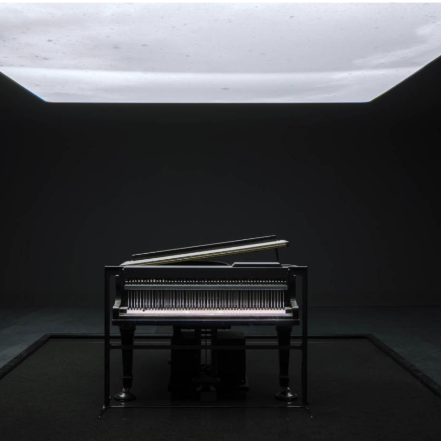 A grand piano with its lid open, positioned in a dark room under a softly illuminated ceiling.